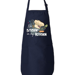 May include: A navy blue kitchen apron with a white pocket and a graphic that says "No Bitchin' in my Kitchen" with a chef's hat, rolling pin, spatula, and whisk.