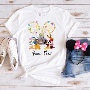 Puede incluir: Camiseta blanca con un gr&aacute;fico de Disney colorido con Mickey y Minnie Mouse, Donald y Daisy Duck, Goofy y un castillo. La camiseta tiene el texto "Your Text" debajo. Zapatillas blancas y pantalones cortos de mezclilla con una diadema de orejas de Minnie Mouse tambi&eacute;n est&aacute;n en la imagen.