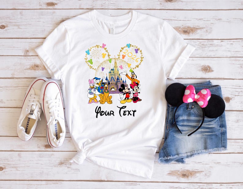 Puede incluir: Camiseta blanca con un gr&aacute;fico de Disney colorido con Mickey y Minnie Mouse, Donald y Daisy Duck, Goofy y un castillo. La camiseta tiene el texto "Your Text" debajo. Zapatillas blancas y pantalones cortos de mezclilla con una diadema de orejas de Minnie Mouse tambi&eacute;n est&aacute;n en la imagen.