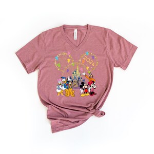 Puede incluir: Camiseta de cuello en V color malva con un gr&aacute;fico colorido de personajes de Disney, incluyendo Mickey y Minnie Mouse, Donald Duck y Goofy, frente a un castillo. El a&ntilde;o "2026" se muestra en oro sobre los personajes.