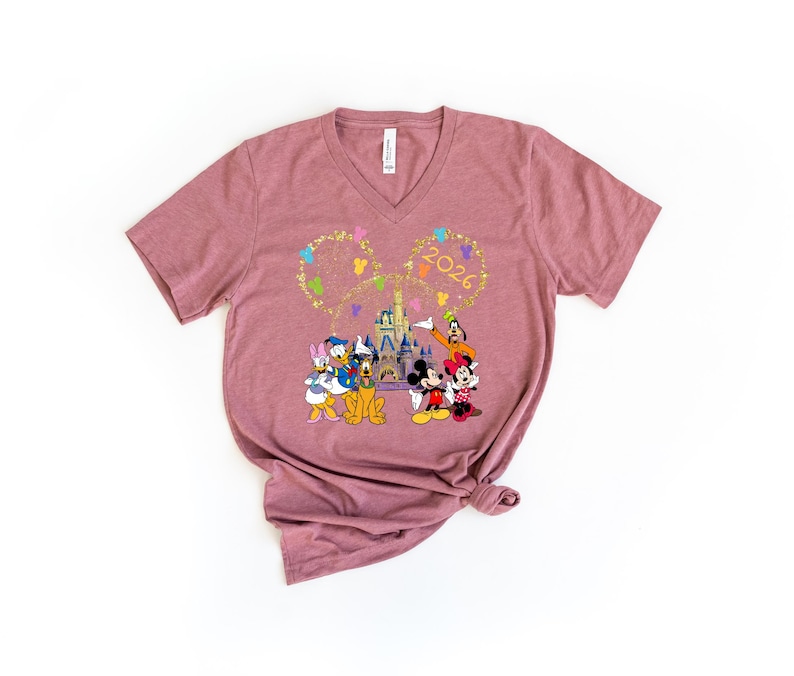 Puede incluir: Camiseta de cuello en V color malva con un gr&aacute;fico colorido de personajes de Disney, incluyendo Mickey y Minnie Mouse, Donald Duck y Goofy, frente a un castillo. El a&ntilde;o "2026" se muestra en oro sobre los personajes.
