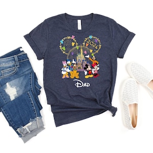 Puede incluir: Camiseta azul marino con un dise&ntilde;o de Disney con Mickey y Minnie Mouse, el Pato Donald y Goofy frente a un castillo. La camiseta tiene el texto "2026" y "Dad". La imagen tambi&eacute;n incluye vaqueros azules y zapatos blancos.