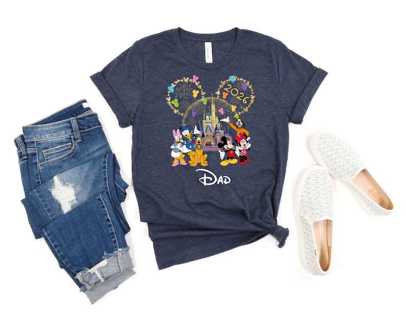 Puede incluir: Camiseta azul marino con un dise&ntilde;o de Disney con Mickey y Minnie Mouse, el Pato Donald y Goofy frente a un castillo. La camiseta tiene el texto "2026" y "Dad". La imagen tambi&eacute;n incluye vaqueros azules y zapatos blancos.