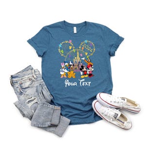Puede incluir: Una camiseta azul con un dise&ntilde;o de Disney con Mickey Mouse, Minnie Mouse, Donald Duck y otros personajes. El dise&ntilde;o incluye el texto "2026" y "Your Text". La camiseta se combina con vaqueros azules y zapatillas blancas.