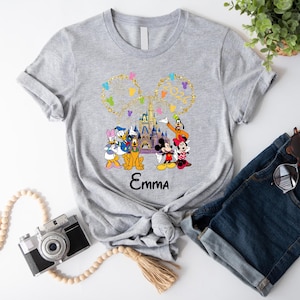 Puede incluir: Camiseta gris con un dise&ntilde;o colorido de personajes de Disney, incluyendo Mickey y Minnie Mouse, Donald Duck y Goofy. El dise&ntilde;o incluye el texto "Emma" y un castillo. Las mangas de la camiseta est&aacute;n dobladas.