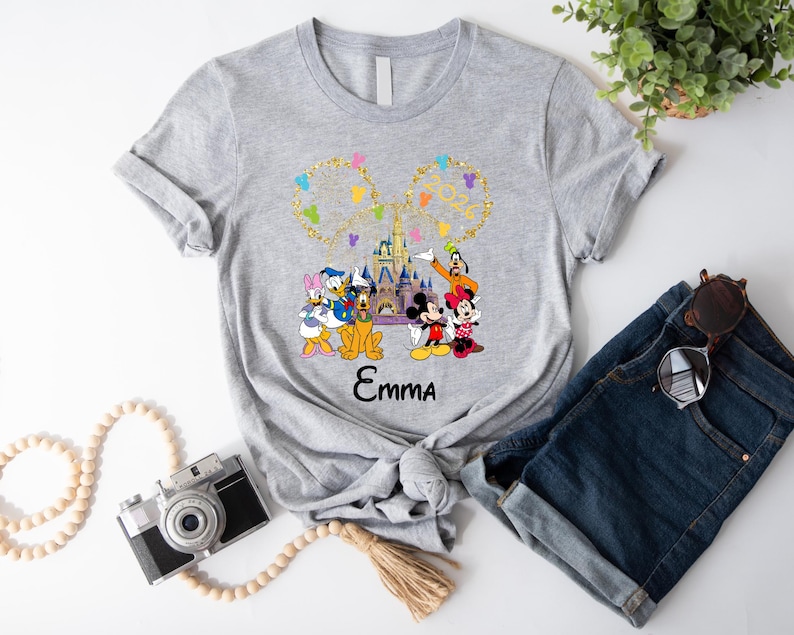Puede incluir: Camiseta gris con un dise&ntilde;o colorido de personajes de Disney, incluyendo Mickey y Minnie Mouse, Donald Duck y Goofy. El dise&ntilde;o incluye el texto "Emma" y un castillo. Las mangas de la camiseta est&aacute;n dobladas.