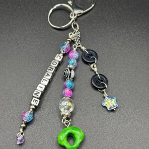 May include: A handmade keychain with silver-tone hardware. The keychain features a beaded charm with the name "CORALINE" spelled out in white letter beads. Additional charms include black buttons, a green donut-shaped charm, and colorful beads.