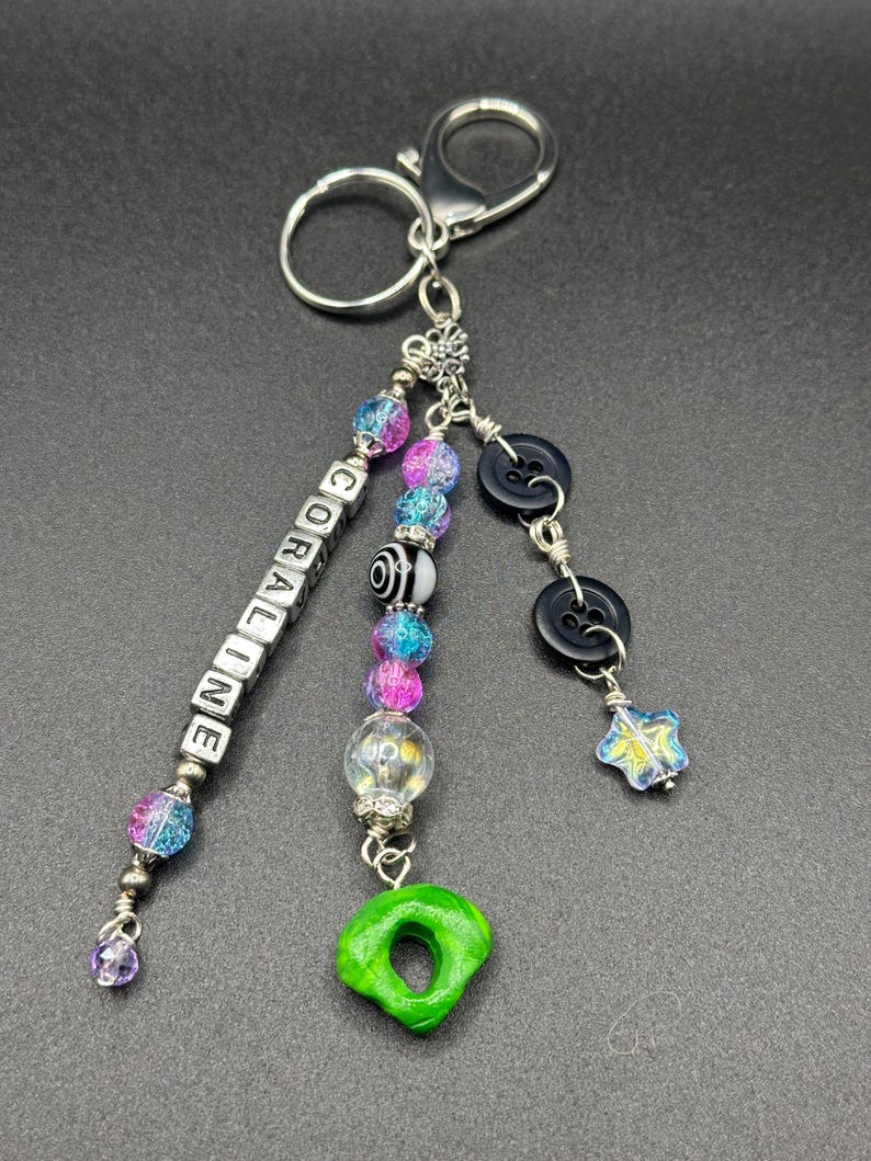 May include: A handmade keychain with silver-tone hardware. The keychain features a beaded charm with the name "CORALINE" spelled out in white letter beads. Additional charms include black buttons, a green donut-shaped charm, and colorful beads.