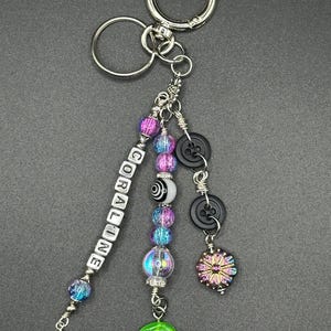 May include: A silver keychain with a variety of decorative charms. The charms include beads in shades of purple and blue, black buttons, a green charm, and a charm that says "CORALINE".