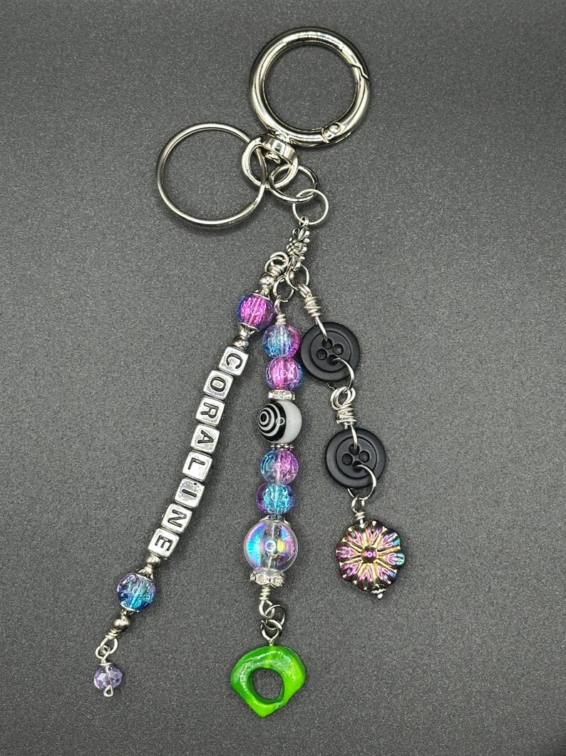 May include: A silver keychain with a variety of decorative charms. The charms include beads in shades of purple and blue, black buttons, a green charm, and a charm that says "CORALINE".