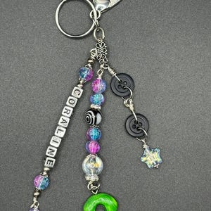 May include: A handmade keychain with a silver clasp and ring. The keychain features beads in shades of blue, purple, and clear, along with black buttons and a green charm. The word "CORALINE" is spelled out in white letter beads.