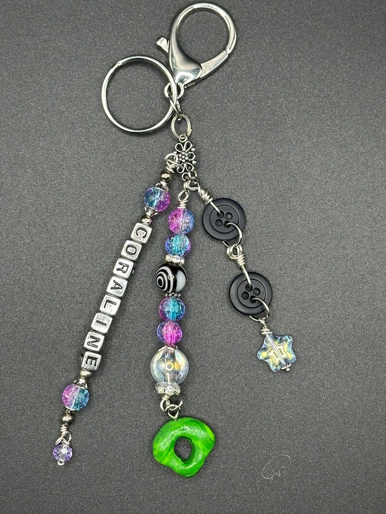 May include: A handmade keychain with a silver clasp and ring. The keychain features beads in shades of blue, purple, and clear, along with black buttons and a green charm. The word "CORALINE" is spelled out in white letter beads.