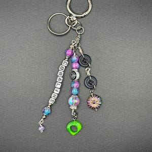 May include: A handmade keychain with a silver keyring and multiple charms. The charms include beads in shades of purple, blue, and clear, black buttons, and a green, eye-shaped charm. The word "CORALINE" is spelled out in white letter beads.