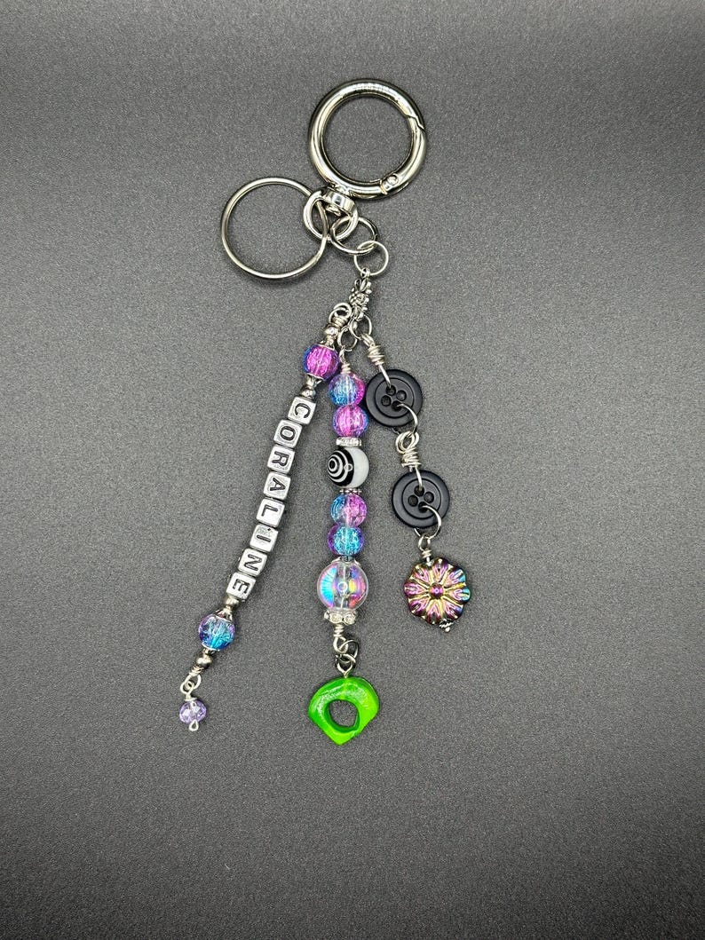 May include: A handmade keychain with a silver keyring and multiple charms. The charms include beads in shades of purple, blue, and clear, black buttons, and a green, eye-shaped charm. The word "CORALINE" is spelled out in white letter beads.