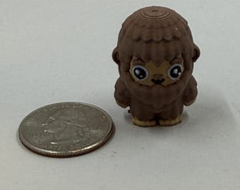 May include: A small, brown figurine of a cartoonish Bigfoot, with large eyes and a furry appearance. The figurine is next to a U.S. quarter for size comparison. The Bigfoot has a textured head and a light brown face and feet.