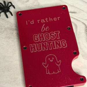 May include: A rectangular red metal wallet featuring the engraved text I'd rather be GHOST HUNTING and a small ghost illustration. The wallet has black screws at the corners and a side cutout for card access, with a black elastic band at the base.