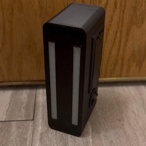 May include: A black rectangular electronic device with two vertical white light strips on the front. It has a red power button on the side. The device is sitting on a gray surface.
