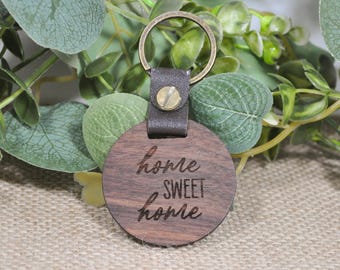 Könnte beinhalten: Ein runder Holzschlüsselanhänger mit einem braunen Lederband und einem Metallring. Der Schlüsselanhänger ist mit dem Text "home sweet home" graviert.