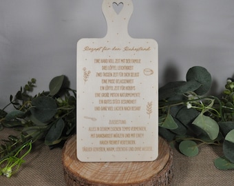May include: A light-colored wooden cutting board with a handle and a heart-shaped cutout. The board has engraved text in German, along with small illustrations. It rests on a wooden base, surrounded by greenery.