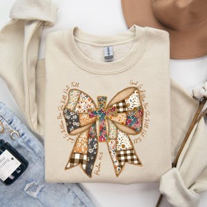 May include: A cream-coloured sweatshirt with a large bow design and a cross in the centre. The bow is made of various floral and plaid patterns. The text "God Is Within Her She Will Not Fall" and "Psalm 46:5" are printed around the bow.