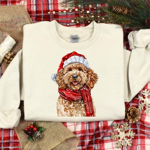 May include: Cream-colored sweatshirt featuring a watercolor illustration of a dog wearing a Santa hat and a red scarf. The dog has brown fur and a happy expression, perfect for the holiday season.