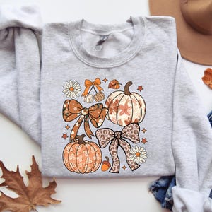 Puede incluir: Una sudadera gris claro con un gráfico de temática otoñal. El gráfico incluye calabazas, lazos, estrellas y flores en tonos naranja, marrón y crema. El diseño tiene una estética vintage y otoñal.