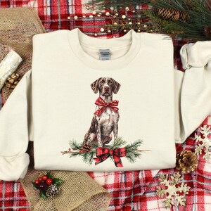 May include: Cream-colored sweatshirt featuring a watercolor illustration of a German Shorthaired Pointer dog wearing a red bow. The dog sits atop a sprig of pine with a red and black plaid bow. A festive holiday design.