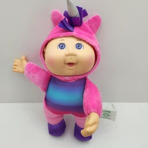 May include: A Cabbage Patch Kids doll dressed in a pink unicorn costume. The doll has blue eyes, a smiling expression, and is wearing a hooded outfit with a purple horn and ears. The body of the doll has a rainbow gradient design.