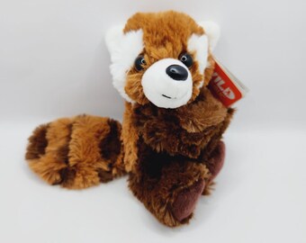 May include: A plush red panda soft toy with brown and white fur. The toy has a black nose and black eyes. The red panda is sitting with its tail curled around its body. The toy has a tag that says "WILD" on it.
