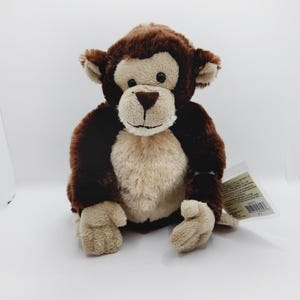 May include: A brown and tan plush monkey toy with a smiling face. The monkey is sitting with its legs crossed and its arms outstretched. The toy has a tag attached to its right arm.