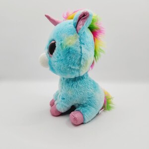 Op de afbeelding: Een blauwe en regenboogkleurige pluche eenhoorn met grote ogen en een roze hoorn. De eenhoorn zit op een witte achtergrond.