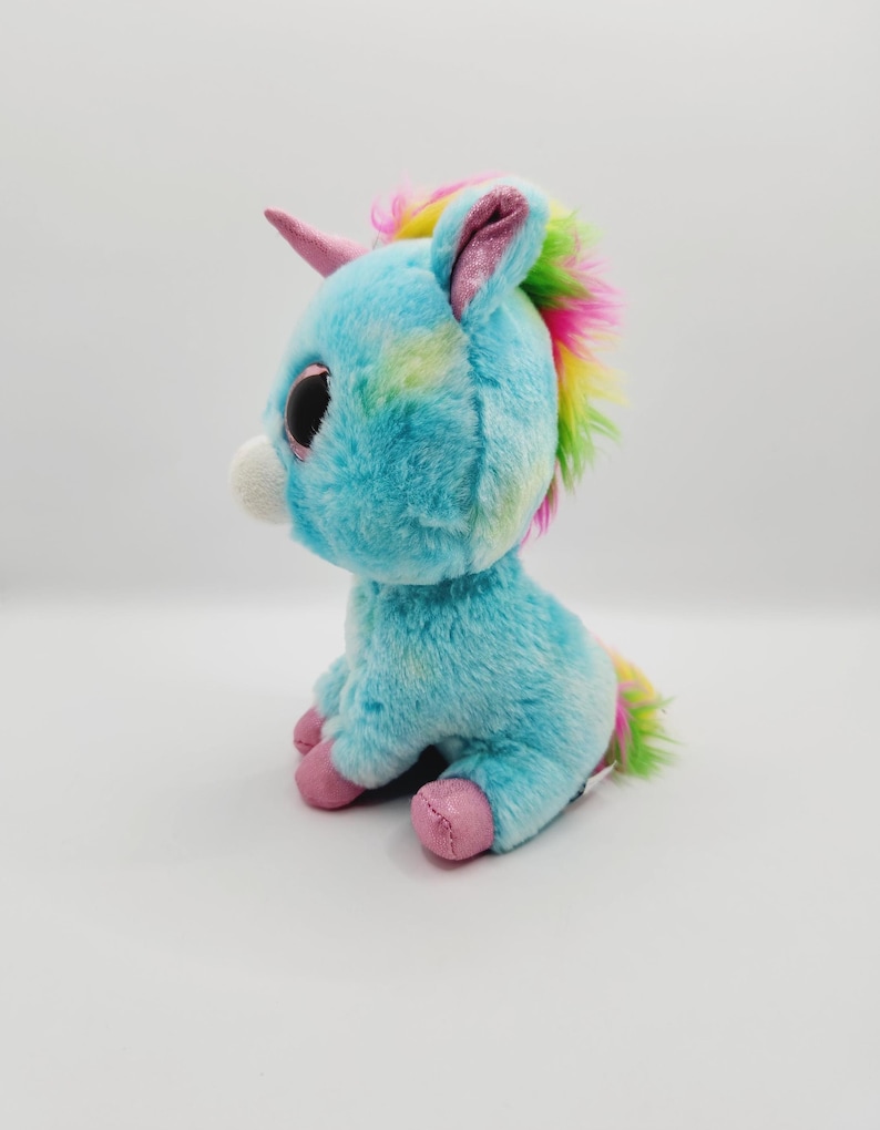 Op de afbeelding: Een blauwe en regenboogkleurige pluche eenhoorn met grote ogen en een roze hoorn. De eenhoorn zit op een witte achtergrond.