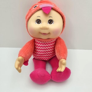 May include: A plush doll with a round face, wearing a coral-colored flamingo-themed outfit. The doll has a pink hat with a beak, a zig-zag patterned shirt, and pink pants. The doll's eyes are brown, and it has rosy cheeks.