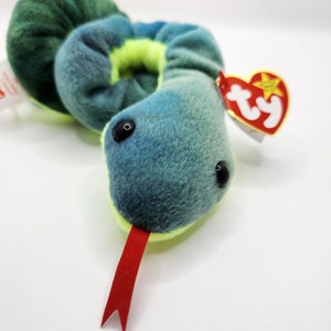 May include: A plush snake toy featuring a soft, fuzzy texture with a gradient of blue and green colours on the top and a bright yellow underside. The snake has black plastic eyes, a red felt tongue, and a heart-shaped Ty Beanie Baby tag attached.