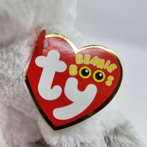 May include: A plush, gray and white stuffed animal with a red heart-shaped tag. The tag has the words "Beanie Boos" in yellow letters and the Ty logo. The stuffed animal is soft and fluffy.