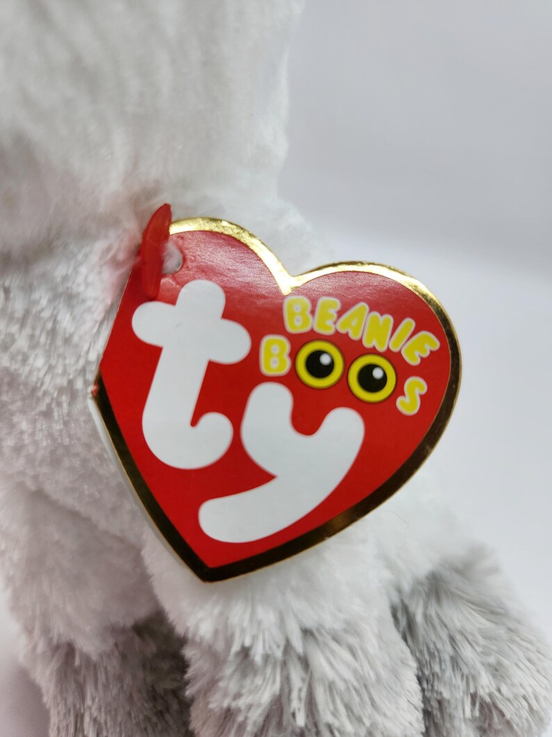 May include: A plush, gray and white stuffed animal with a red heart-shaped tag. The tag has the words "Beanie Boos" in yellow letters and the Ty logo. The stuffed animal is soft and fluffy.