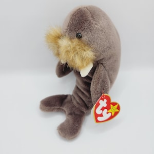May include: A brown plush walrus with a brown and white face and a brown and white mustache. The walrus is wearing a red heart-shaped tag with the word "Ty" on it.