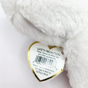 May include: A close-up of a white plush toy with a heart-shaped tag. The tag has the text "SAFETY PRECAUTION" and "Retain Tag For Reference". The toy is soft and fluffy.