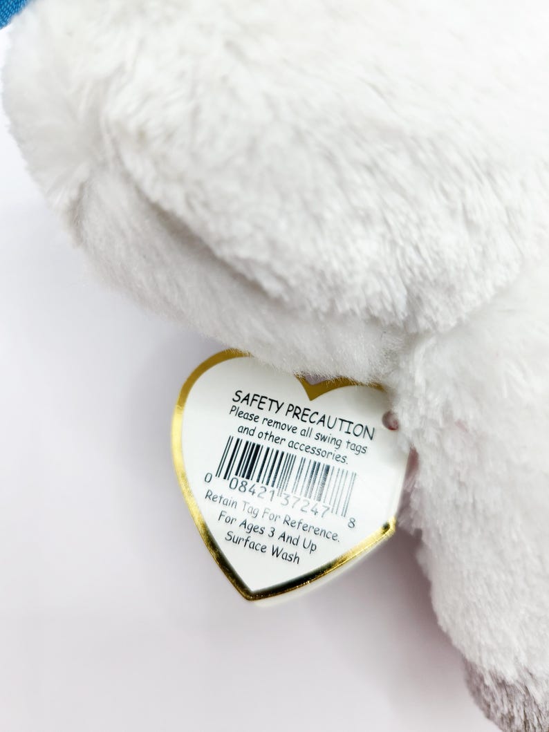 May include: A close-up of a white plush toy with a heart-shaped tag. The tag has the text "SAFETY PRECAUTION" and "Retain Tag For Reference". The toy is soft and fluffy.
