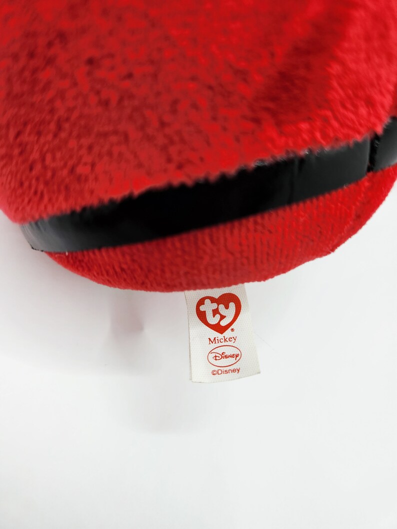 May include: Red plush Mickey Mouse toy with a black band around the middle. The toy has a white Ty tag with the words "Mickey" and "Disney" on it.