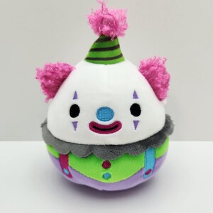 Può includere: Bambola pagliaccio di peluche con testa bianca, occhi neri e naso blu. Il pagliaccio ha un cappello a righe verdi e nere, capelli rosa e un corpo colorato con accenti verdi, viola e grigi. Un giocattolo divertente e stravagante.