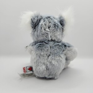 May include: A gray and white plush koala bear sitting on a white surface. The bear has a white tag on its foot that says "Ganz" with a red heart.