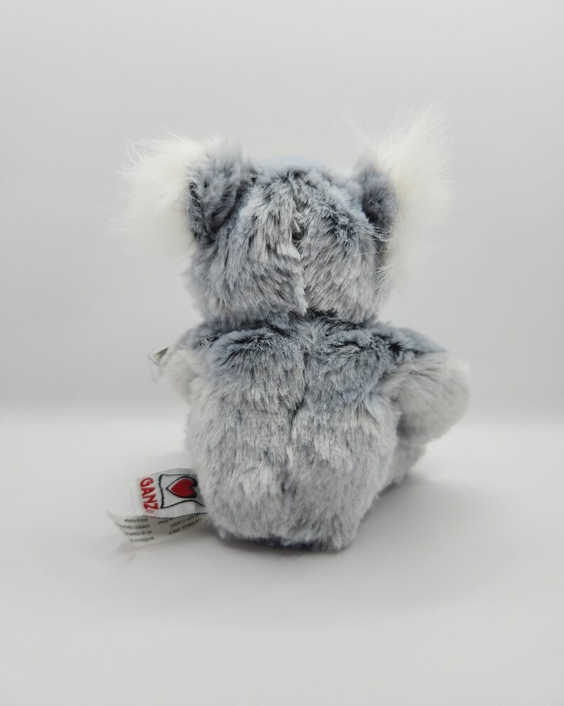 May include: A gray and white plush koala bear sitting on a white surface. The bear has a white tag on its foot that says "Ganz" with a red heart.
