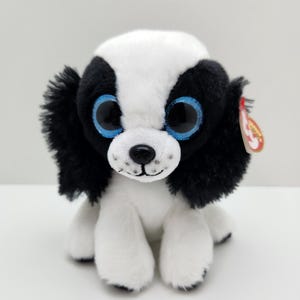 May include: A soft toy dog with a black and white coat, large blue eyes, and a black nose. The dog has floppy black ears and is sitting upright. A red tag is attached to the ear.