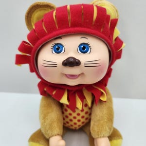 May include: A plush doll dressed as a lion, featuring a red and yellow felt mane and head covering. The doll has a tan body with a red polka-dotted belly, blue eyes, and a smiling expression. The doll is sitting.