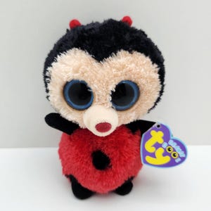 Può includere: Un peluche a forma di coccinella con corpo rotondo rosso a pois neri, testa pelosa nera e beige, grandi occhi blu scintillanti e due piccole antenne rosse. Un'etichetta viola Ty Beanie Boos a forma di cuore è attaccata al lato.