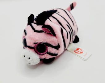 Puede incluir: Un pequeño peluche de cebra con pelaje rosa suave y rayas negras. Presenta grandes ojos redondos de color rosa brillante, un hocico blanco con puntos negros bordados y una cola pequeña. Tiene una etiqueta de tela blanca con el logo rojo de Ty en forma de corazón.