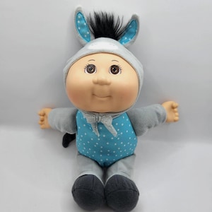 May include: A doll with a round face, brown eyes, and black hair, wearing a grey donkey-themed hooded onesie. The hood features upright ears with blue and white polka dot fabric inside. The onesie has a blue polka dot torso and grey limbs.