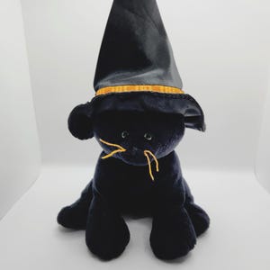 Puede incluir: Un juguete de peluche negro con forma de gato que lleva un sombrero de bruja negro con una banda dorada. El gato tiene ojos verdes y bigotes amarillos. El juguete est&aacute; sobre una superficie blanca.