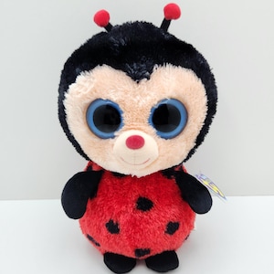 Può includere: Un peluche a forma di coccinella con un corpo rotondo e soffice rosso a pois neri e una testa nera. Presenta grandi occhi blu lucidi, un piccolo naso rosa e due antenne con punte rosse. Il giocattolo ha braccia e piedi di colore nero.
